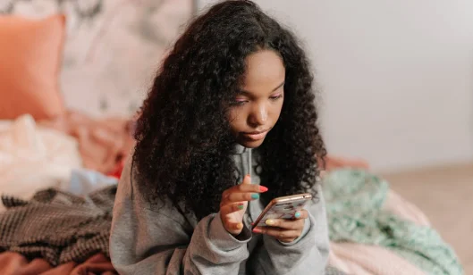Uma adolescente usando um celular e deitada na mesa do quarto.